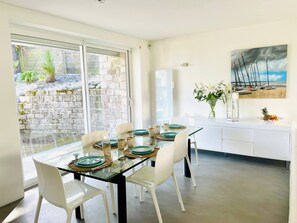 A modern dining area with a glass table, white chairs, and a view of a garden through large windows.