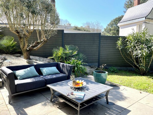 Un patio con un sofá azul oscuro, una mesa de centro de madera con frutas y una revista, y una planta verde en maceta.