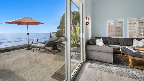 Een woonkamer met een grote schuifpui die uitkomt op een terras met een rode parasol, een grijze bank en uitzicht op zee.
