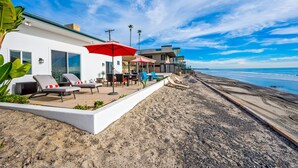 Een strandpand met een patio, uitgerust met ligbedden en parasols, een wit gebouw met grote ramen en uitzicht op de oceaan.