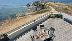 A deck with a table set for dining, overlooking a coastal landscape with rocky cliffs and the sea. - L'écrin marin : Vue exceptionnelle sur l'Océan (Batz-sur-Mer)
