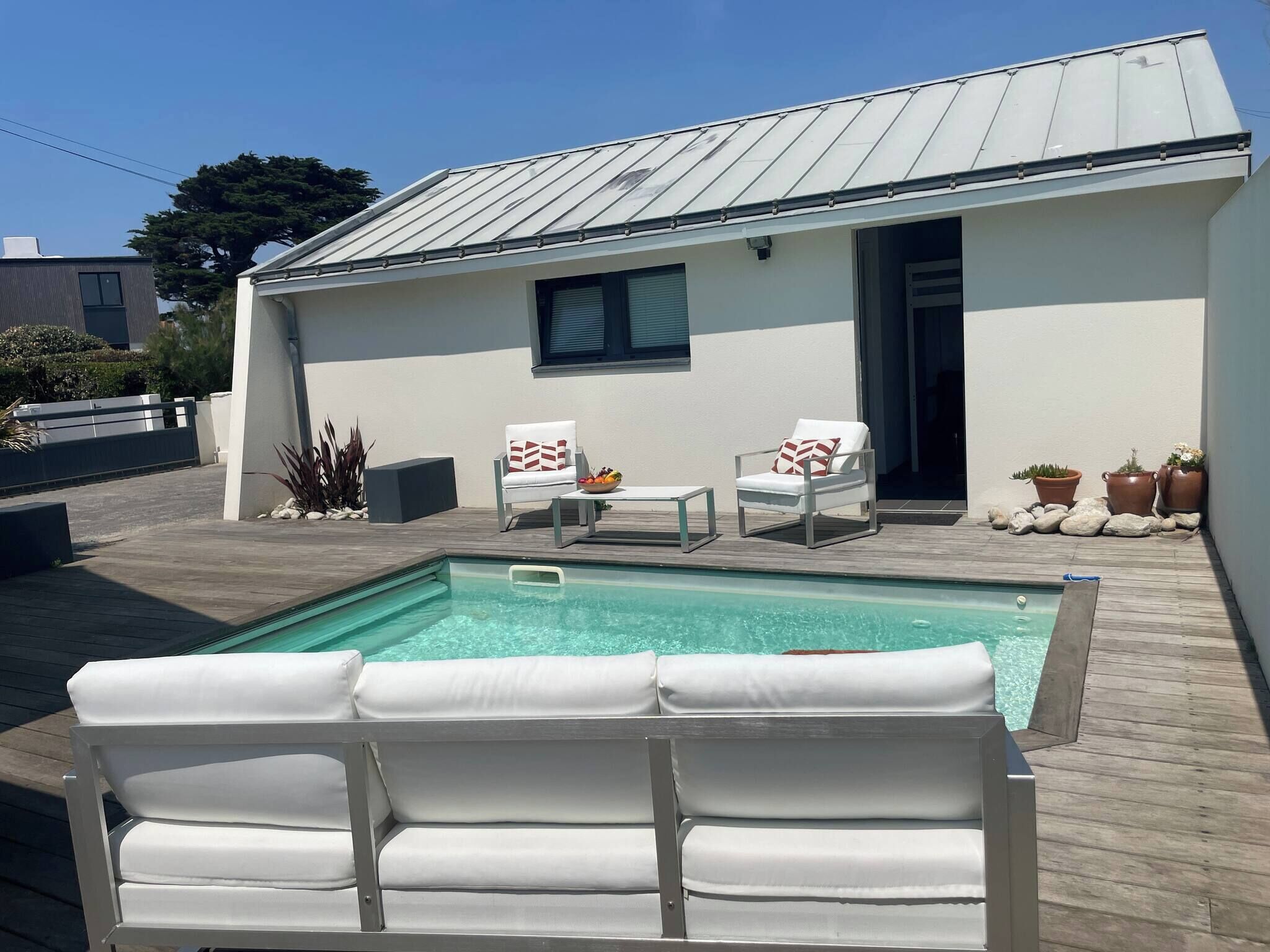 Un espace aménagé au bord de la piscine, comprenant une piscine, deux chaises blanches avec des coussins à motifs et des plantes en pot.