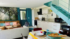 Una sala de estar moderna con un sofá, una mesa de comedor con mantel a rayas y una cocina con armarios blancos y una escalera verde azulada.
