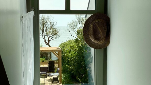 Una ventana con vista a un patio, un sombrero colgado del marco y una planta en maceta en el alféizar.