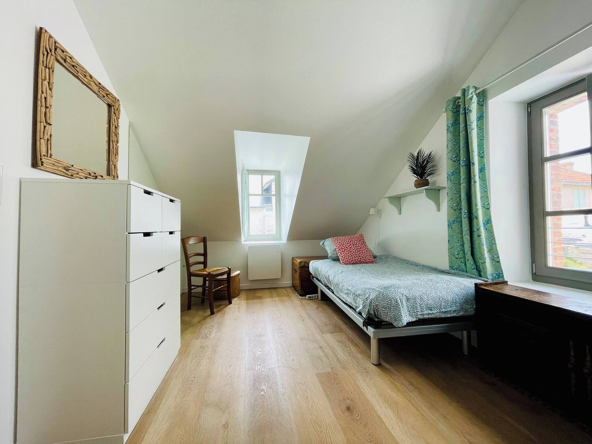 A room with a sloped ceiling, a bed, a chair, a dresser, a window with curtains, and a radiator.