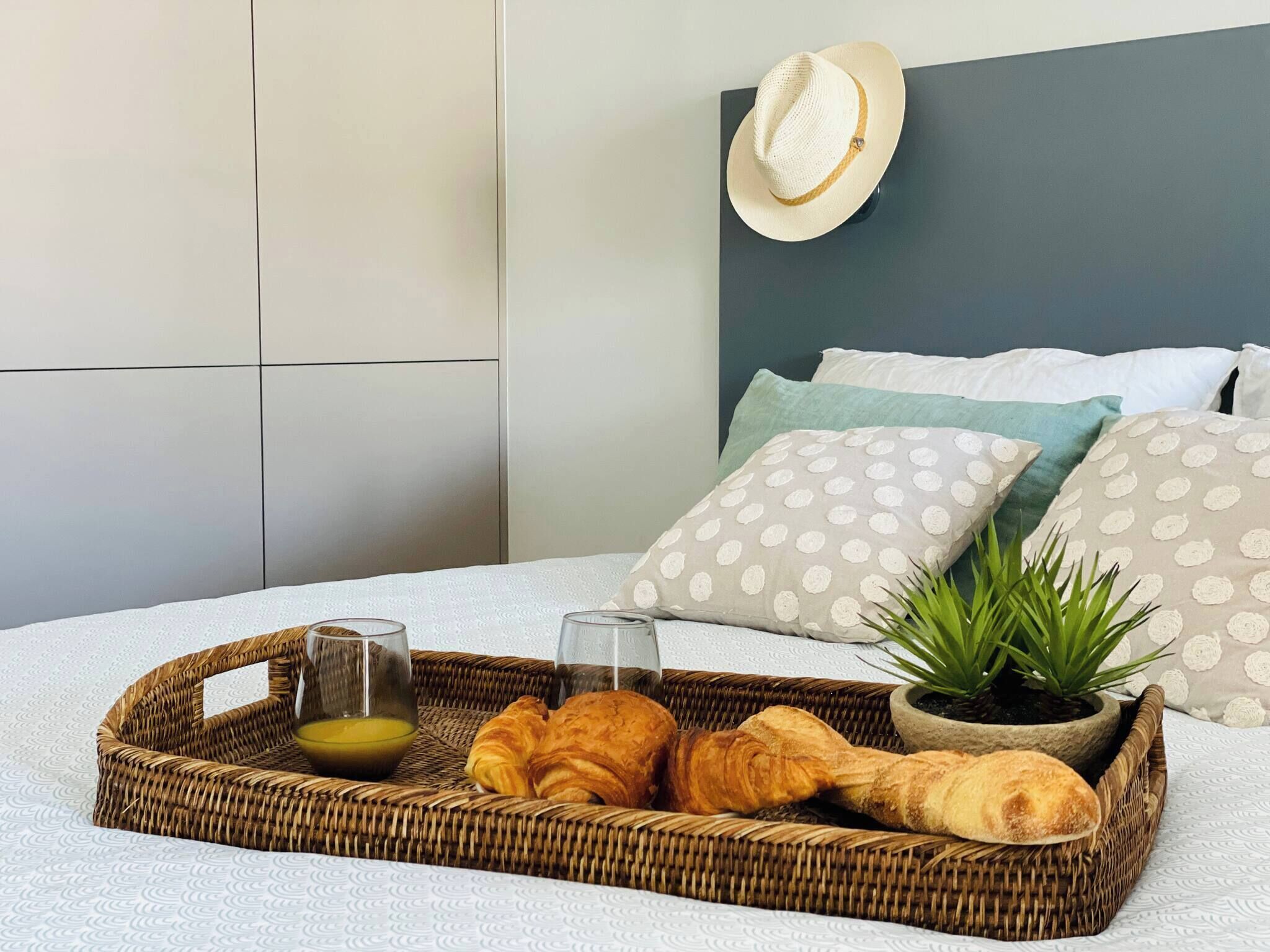 Una cama con una colcha blanca texturizada, una bandeja de mimbre con croissants y un vaso de jugo de naranja, una planta en maceta y un sombrero en la pared.