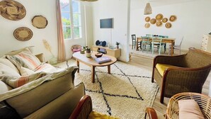 Una sala de estar con sofá, sillas, una mesa de centro, una mesa de comedor con sillas, televisión y adornos en la pared.