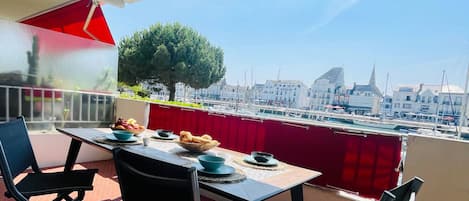 A balcony with a table set for two, chairs, and a view of a marina with boats and buildings.