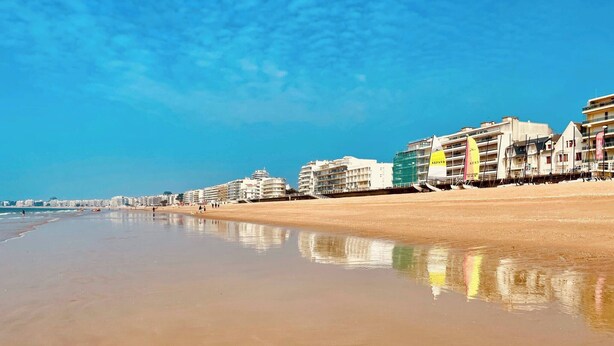 Uma praia com edifícios ao longo da costa, céu claro e água calma refletindo os edifícios.