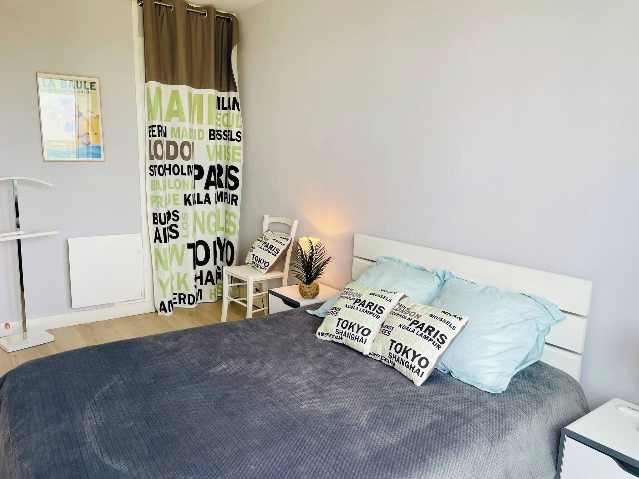 Una camera da letto con un letto, cuscini, una sedia, un tavolino, una pianta e una tenda con nomi di città.