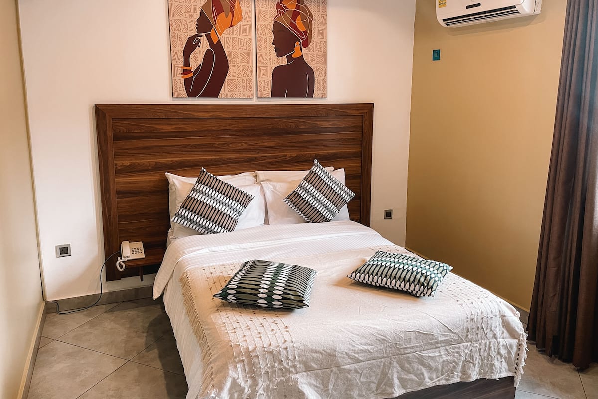 Un dormitorio con cabecera de madera, colcha blanca y almohadas estampadas. Hay dos aires acondicionados instalados en la pared y un teléfono sobre una estantería.