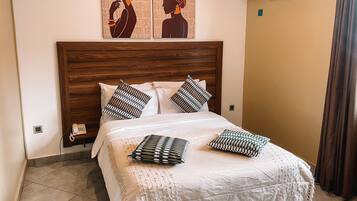 Une chambre à coucher avec une tête de lit en bois, un couvre-lit blanc et des coussins à motifs. On y trouve deux climatiseurs fixés au mur et un téléphone posé sur une étagère.
