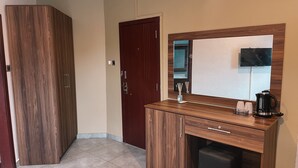 Hotellihuoneessa on puinen vaatekaappi, tumma puinen työpöytä, jossa on peili, musta kahvinkeitin ja pieni musta jääkaappi.