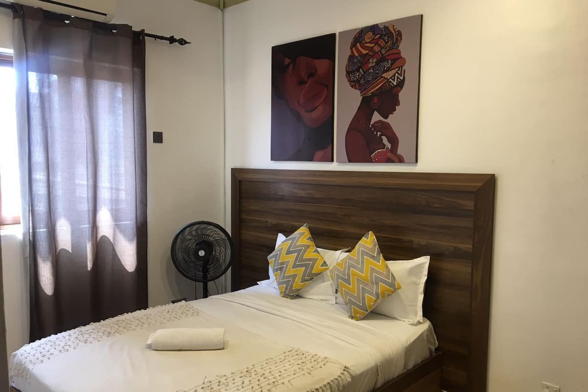Una cama bien hecha con almohadas estampadas, un ventilador de pie y una obra de arte en la pared.