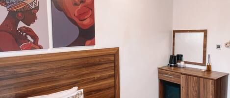 A bedroom with a wooden headboard, a bed with a floral pillow, a nightstand with a coffee maker, and a mirror on the wall.