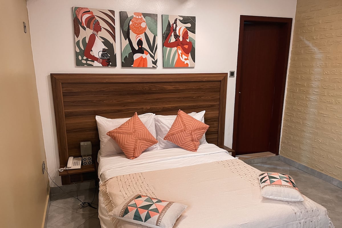 Una cama bien hecha con almohadones estampados, un cabecero de madera y una pared con tres obras de arte.