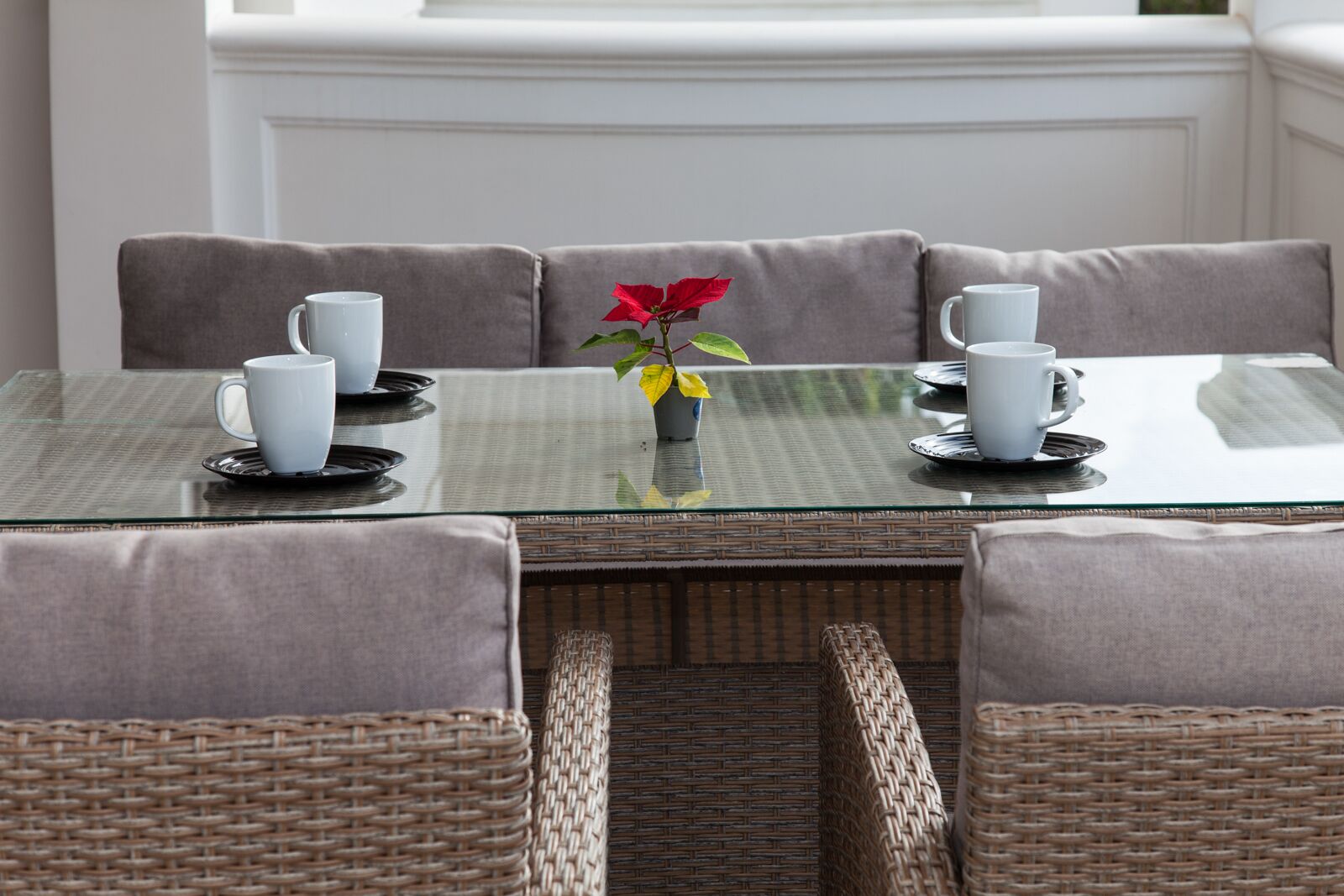 A dining table with four white cups and saucers, a vase with a red flower, and wicker chairs.