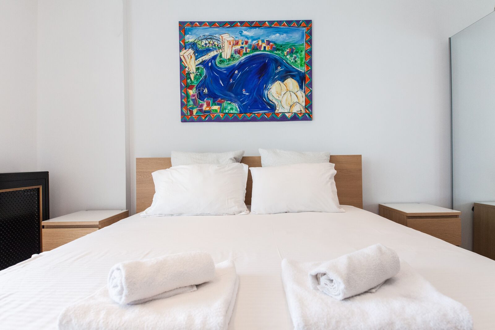 Een hotelkamer met een groot bed, twee witte kussens, een houten hoofdbord en een kleurrijk schilderij aan de muur.