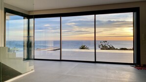 Ein geräumiger Raum mit großen, vom Boden bis zur Decke reichenden Fenstern, der einen Blick auf das Meer und einen Sonnenuntergang bietet.