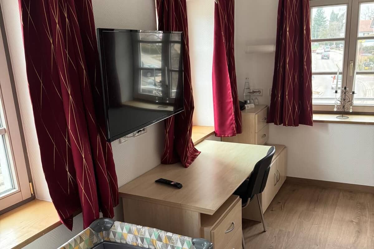 Une chambre avec un lit, un bureau, une télévision et une fenêtre avec des rideaux.