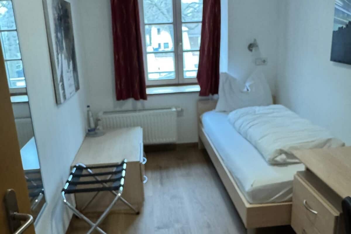 Une petite pièce propre, avec un lit, une table de chevet, un radiateur, une fenêtre avec des rideaux et un tableau accroché au mur.