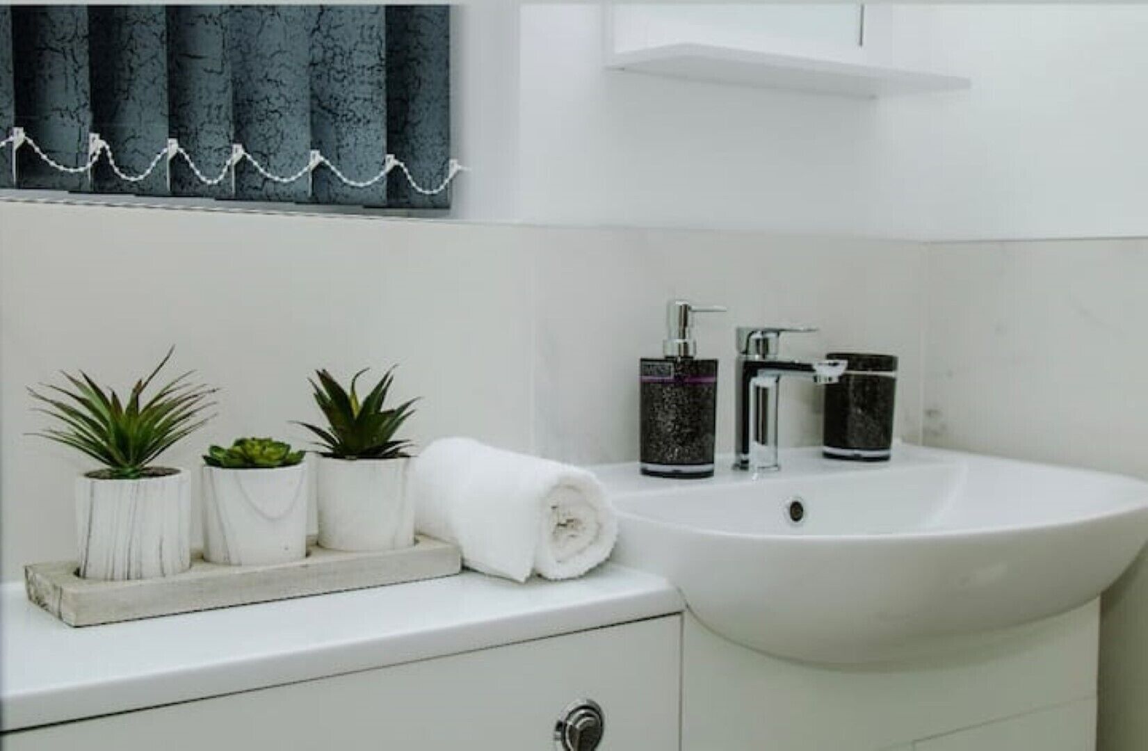 Un bagno con un lavandino bianco, due piante in vaso, un asciugamano e uno specchio con tenda blu.