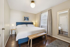A bedroom with a bed, bedside table, lamp, bench, window with curtains, and a mirror.