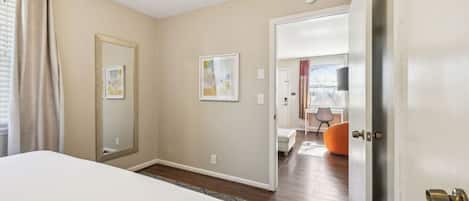A bedroom with a bed, a door leading to a living area, and a view of a cityscape through the window.