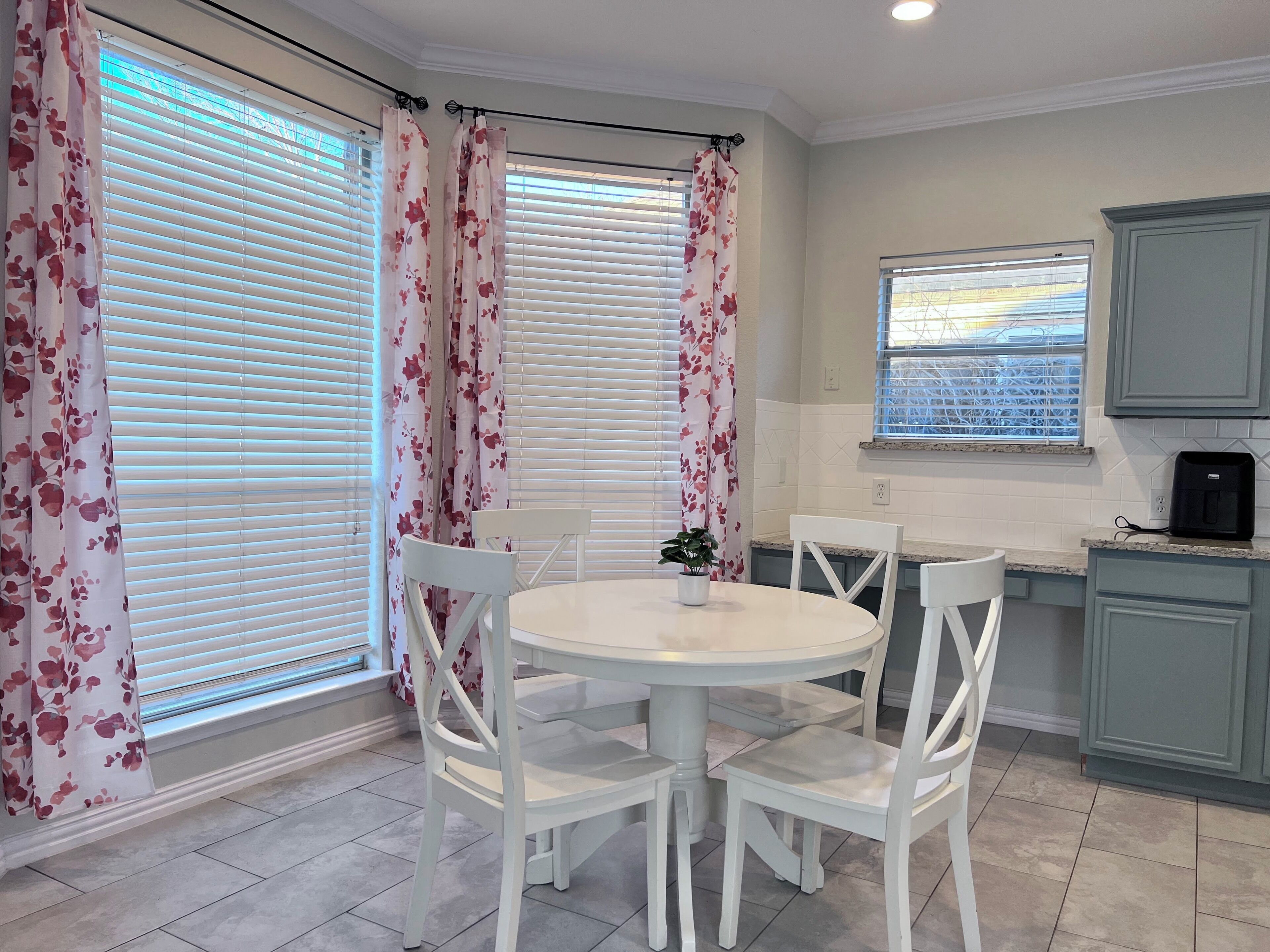 A dining area with a round table and four white chairs, a small potted plant, and a kitchen area with cabinets and a window.