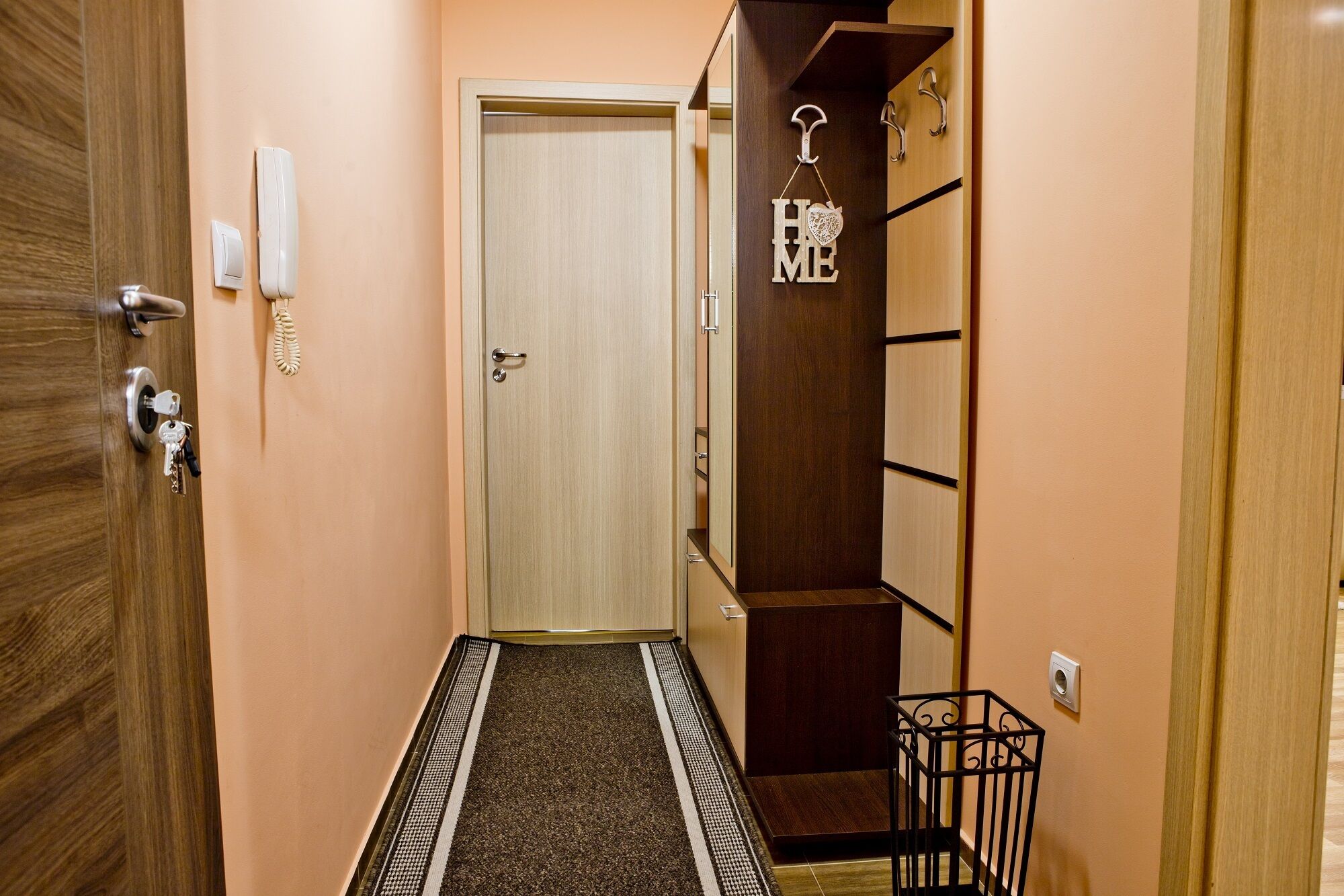Un pasillo con puerta de madera, un interruptor de luz, un perchero para llaves y un pequeño cubo de basura negro.