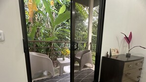 Una stanza con una porta scorrevole in vetro che dà su un balcone con sedie in vimini e un piccolo tavolo, un comò in legno scuro e un vaso con una pianta decorativa.