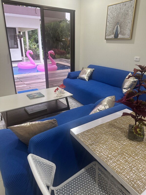 Un soggiorno moderno con un divano blu, un tavolino da caffè bianco e vista su una piscina con fenicotteri rosa.