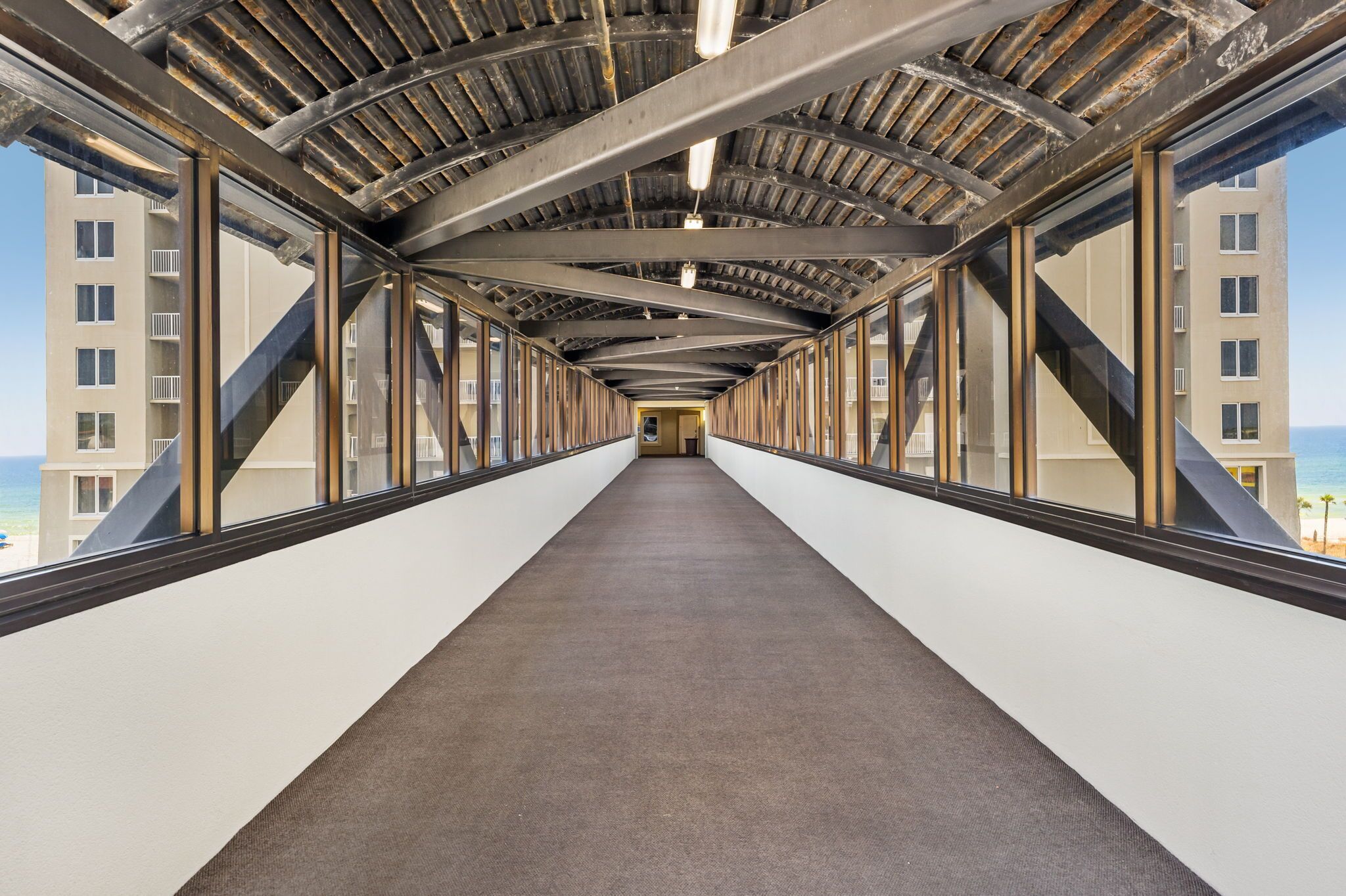 A corridor with a metal roof, large windows, and a carpeted floor, leading to a building with multiple floors and balconies.