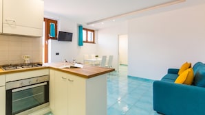 A modern kitchen with white cabinets, a built-in oven, and a wooden countertop. There is a dining area with a white table and chairs, and a blue sofa with yellow cushions. - Nisida View Apartment (Pozzuoli)