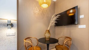 A modern room with a round black table, two wooden chairs, a decorative vase with pampas grass, a wall-mounted TV, and a textured wall. - Sweett | Madeleine VI (Paris)