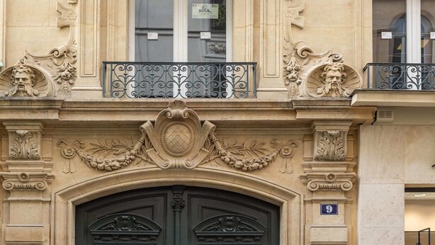 A building facade with ornate architectural details, including a balcony with wrought iron railings and a large, dark-colored entrance door. - Sweett | Madeleine VI (Paris)