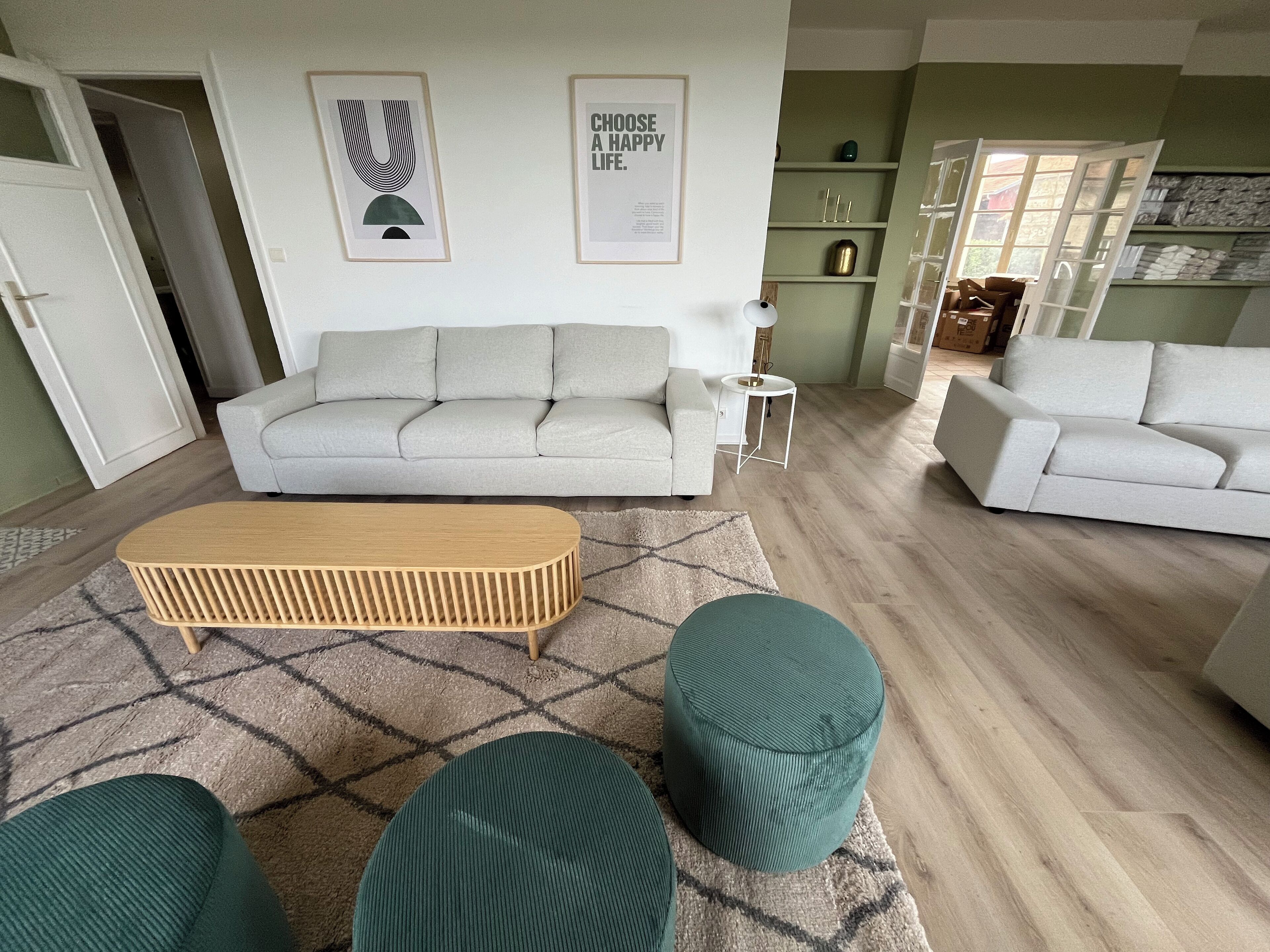Een moderne woonkamer met een bank, poefs en een salontafel. Er hangen ingelijste foto's aan de muur en in de achtergrond staat een boekenplank.