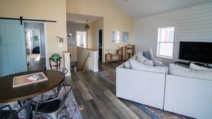 Uma sala de estar espaçosa com um sofá branco, uma mesa de jantar com cadeiras, uma televisão e uma cozinha ao fundo.