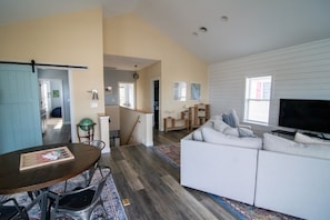 Een ruime woonkamer met een witte bank, een eettafel met stoelen, een televisie en een keuken op de achtergrond.