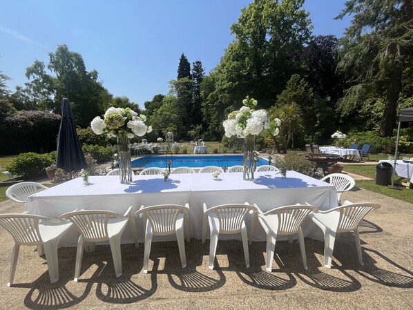 An outdoor dining area with a pool in the background, surrounded by greenery and trees.