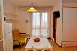 A compact living space with a kitchenette, a dining table with chairs, a colorful sofa, a television mounted on the wall, and a window with sheer curtains. - Relaxing Studio Apartment With Side sea View (San Michele al Tagliamento)