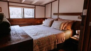Una camera da letto tradizionale in stile giapponese, con pavimento in tatami, un letto basso in legno con biancheria a motivi e un poggiatesta in legno, e una finestra con vista sugli alberi.