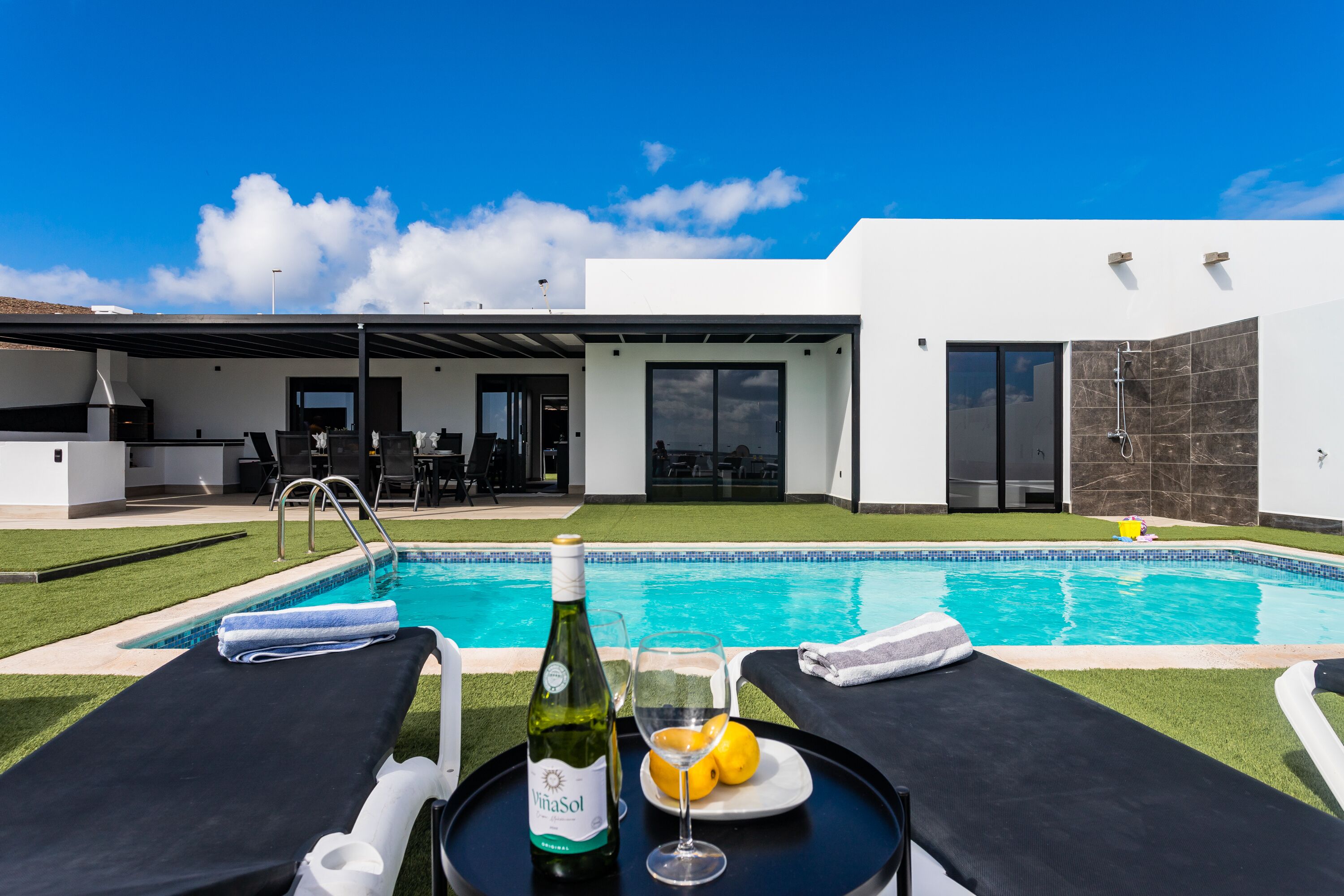 Un área de piscina moderna con sillones de descanso, una botella de vino y un vaso de jugo de naranja en una bandeja.