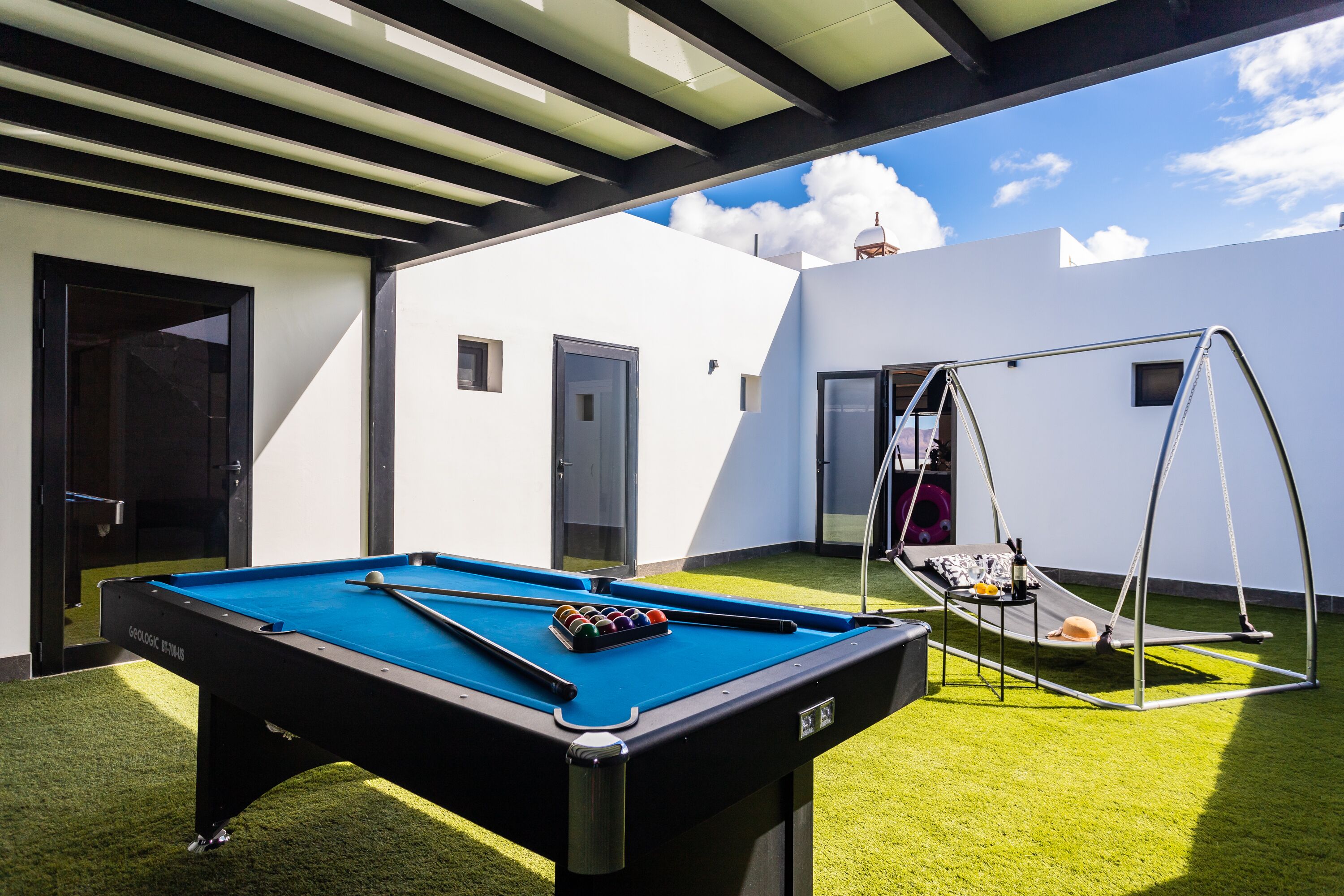 A pool table in a covered outdoor area with a swing chair and a small table with drinks.