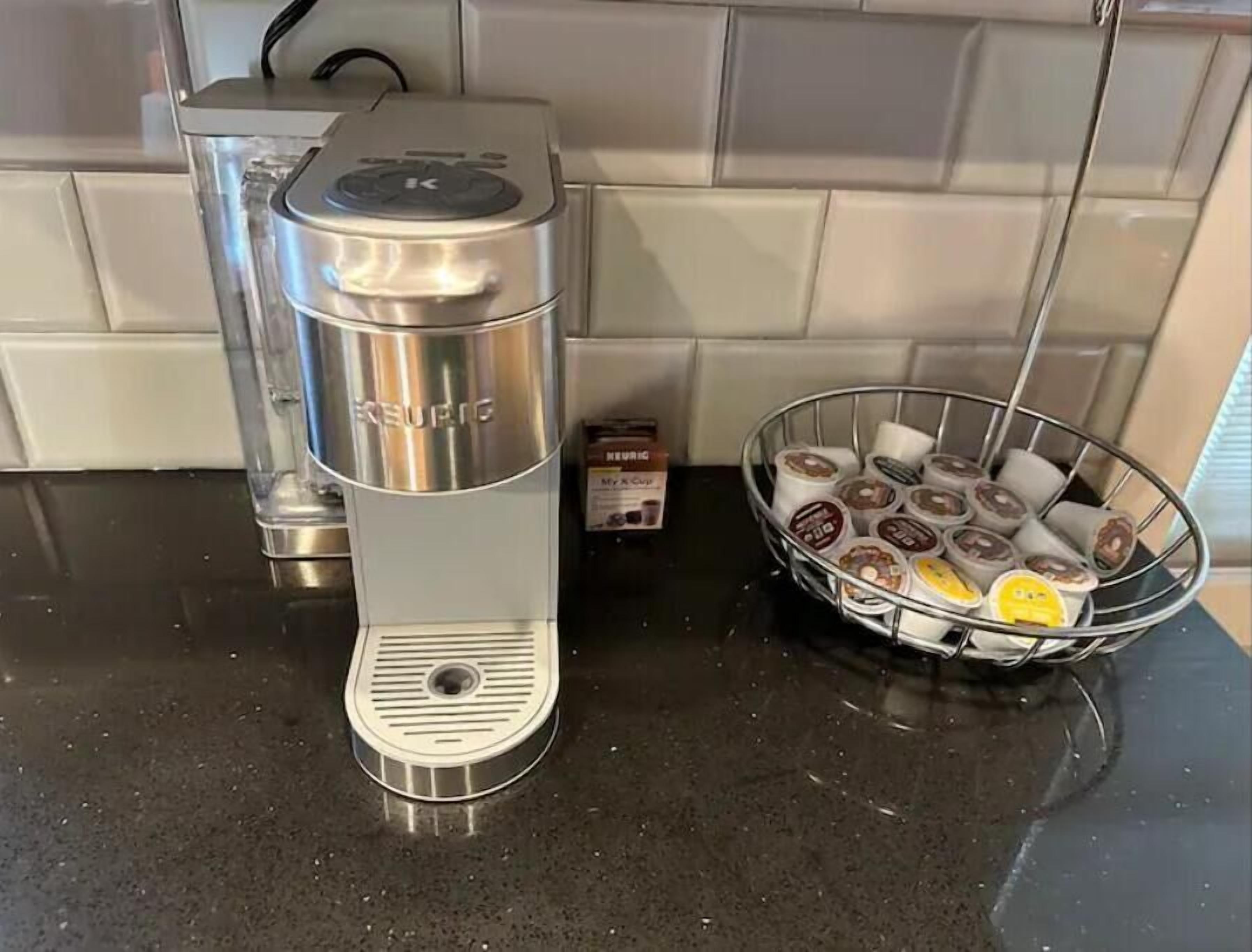Una cafetera Keurig sobre una encimera de cocina, con un cesto de cápsulas de café y una caja de cápsulas reutilizables Keurig My K-Cup.
