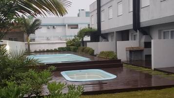 Um pátio com duas hidromassagens, rodeado por vegetação exuberante e um edifício moderno com janelas amplas e uma chaminé.