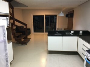 Una cocina con una isla central, armarios blancos y encimera negra. Se ve una escalera que sube a otro nivel, y una puerta corrediza de vidrio al fondo.