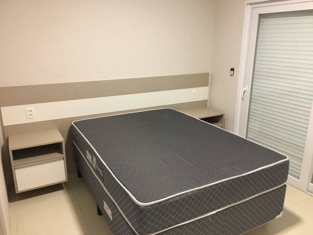 Quarto pequeno com uma cama de solteiro, colchão cinza, mesa de cabeceira e porta fechada.