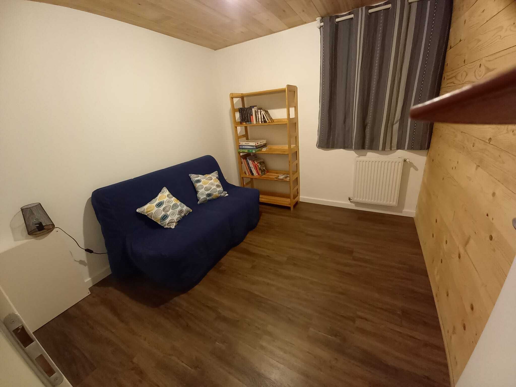 Una habitación pequeña y acogedora con un sofá azul, dos cojines estampados, una estantería de madera y un radiador.