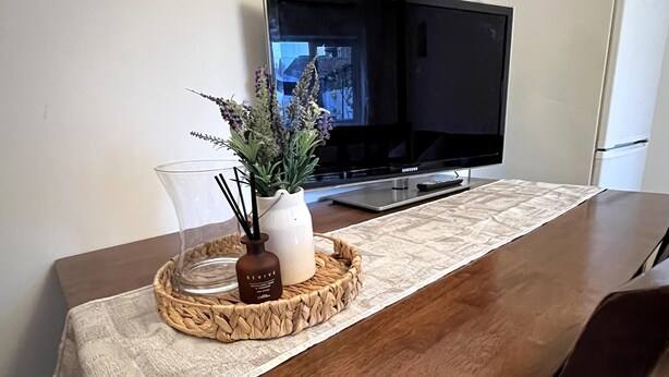 Ein Holztisch mit einer gewebten Tischmatte, eine klare Glasvase mit einer Pflanze und ein weißer Kerzenständer. Ein Flachbildfernseher ist an der Wand über dem Tisch angebracht.